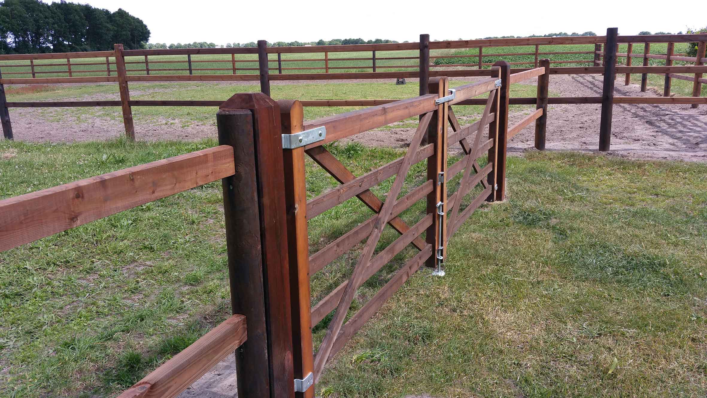 Ein Pferdezaun trennt zwei Paddocks voneinander. Ein doppeltes Holztor ermöglicht den Zugang zu einem Paddock.